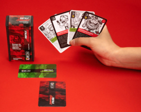 Eine Hand hält Karten in die Kamera, eine Spielschachtel steht auf dem Tisch