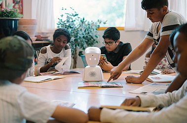 Kinder sitzen an einem Tisch, in der Mitte steht ein weißer Roboter, ein Kind zeigt auf ein Buch, das vor dem Roboter liegt